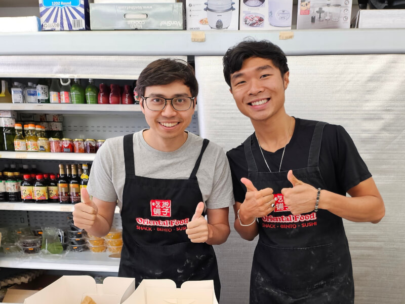 Two men stand behind a table covered in mochi and other desserts, giving a thumbs up